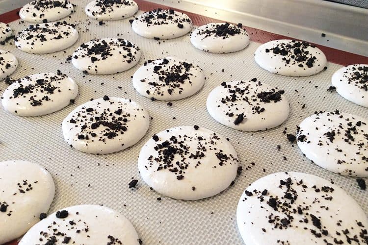 cookies n' cream macarons piped onto silicone mat with cookie crumbs on top
