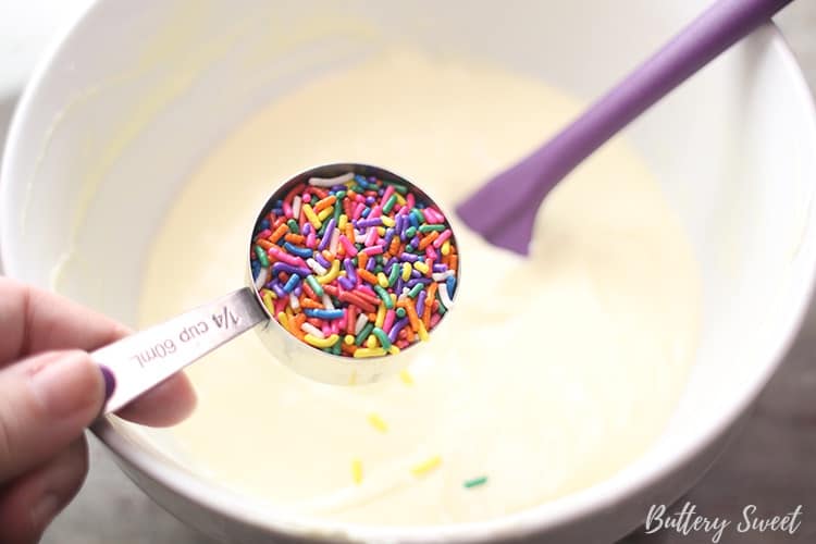 a measuring cup filled with colorful jimmies sprinkles over the bowl of cream cheese mixture